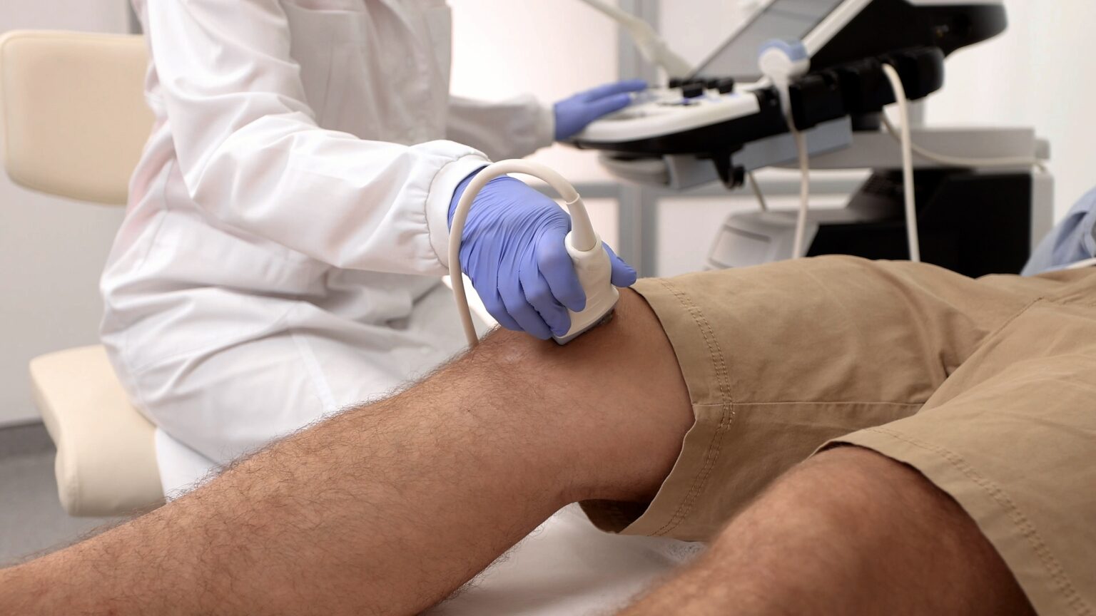 Ultraschallkopf am Knie eines Patienten, im Hintergrund das Sonografiegerät